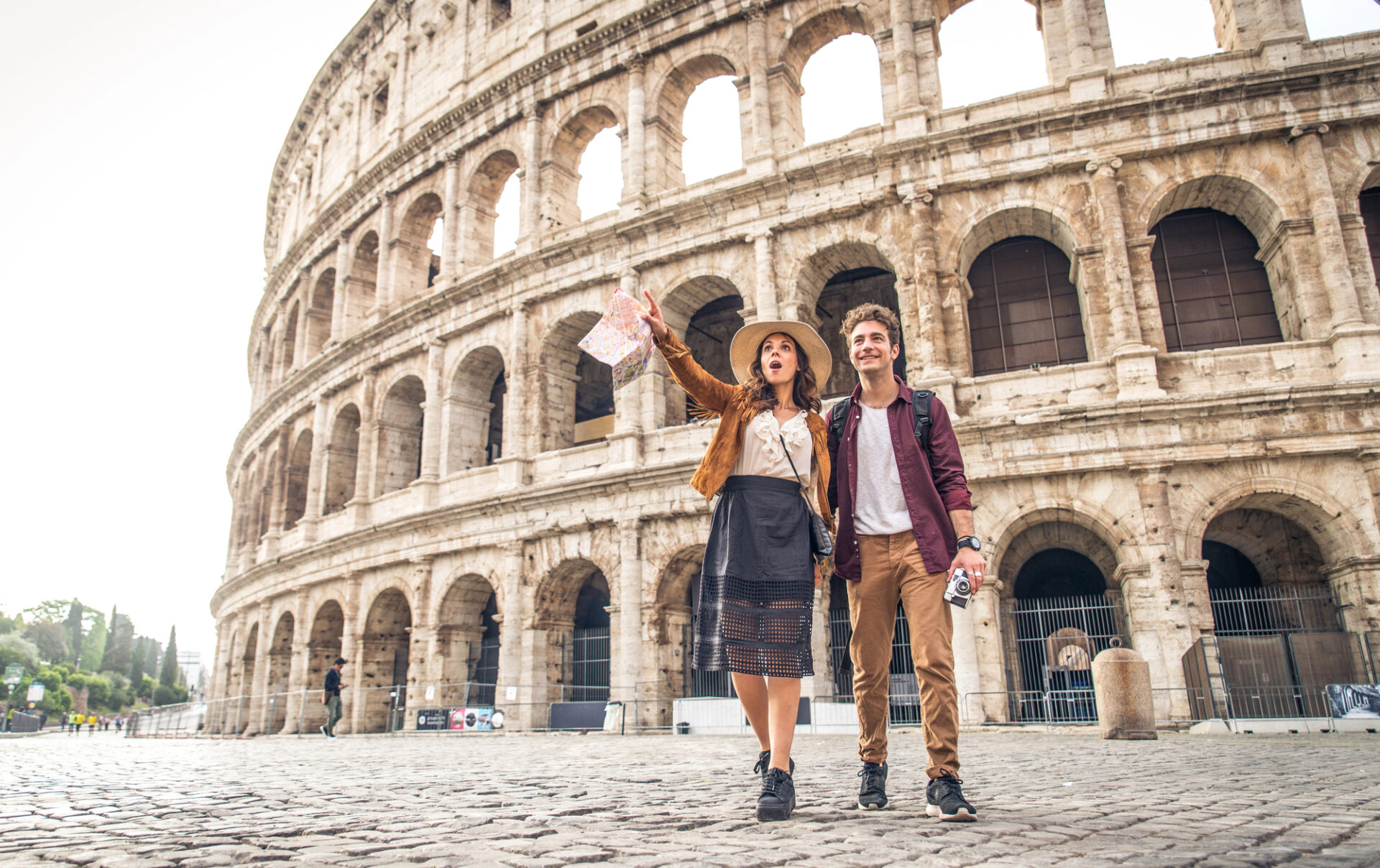 Een toeristenkoppel staat voor het Colosseum in Rome. De vrouw wijst enthousiast naar de lucht terwijl ze een kaart vasthoudt. De man glimlacht terwijl hij naast haar loopt en een camera vasthoudt. Het Colosseum torent hoog boven hen uit.