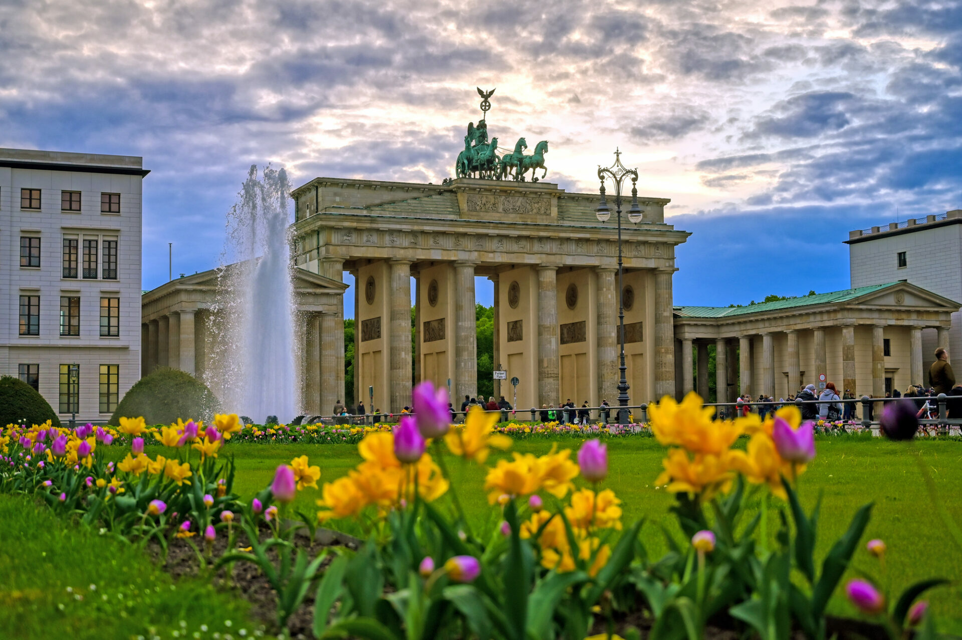 De Brandenburger Tor in Berlijn, Duitsland. Op de voorgrond staan gele en paarse tulpen in het gras, en links van de poort spuit een grote fontein onder een bewolkte hemel.