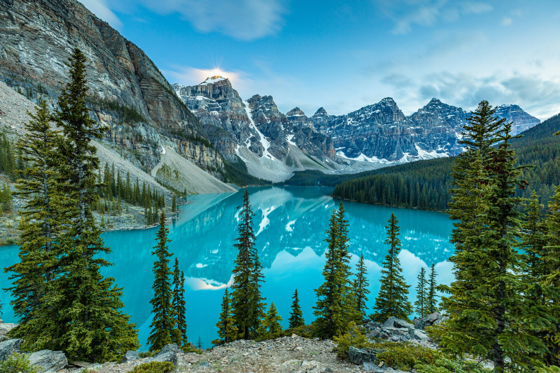 Een adembenemend landschap van Moraine Lake in Canada, met zijn opvallend turquoise water dat de ruige, met sneeuw bedekte bergtoppen van de Valley of the Ten Peaks perfect weerspiegelt. Op de voorgrond staan hoge, groene pijnbomen op een rotsachtige helling die uitkijkt over het meer.